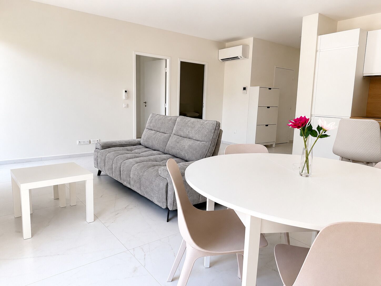 Een moderne woonkamer met een grijze bank, een witte eettafel en een vaas met bloemen.