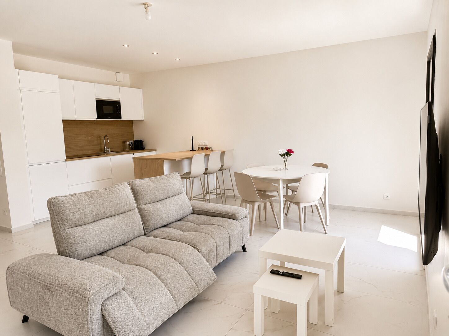 Un salon moderne avec un canapé gris, une petite table blanche et un coin cuisine doté de meubles blancs et d’un plan de travail en bois.