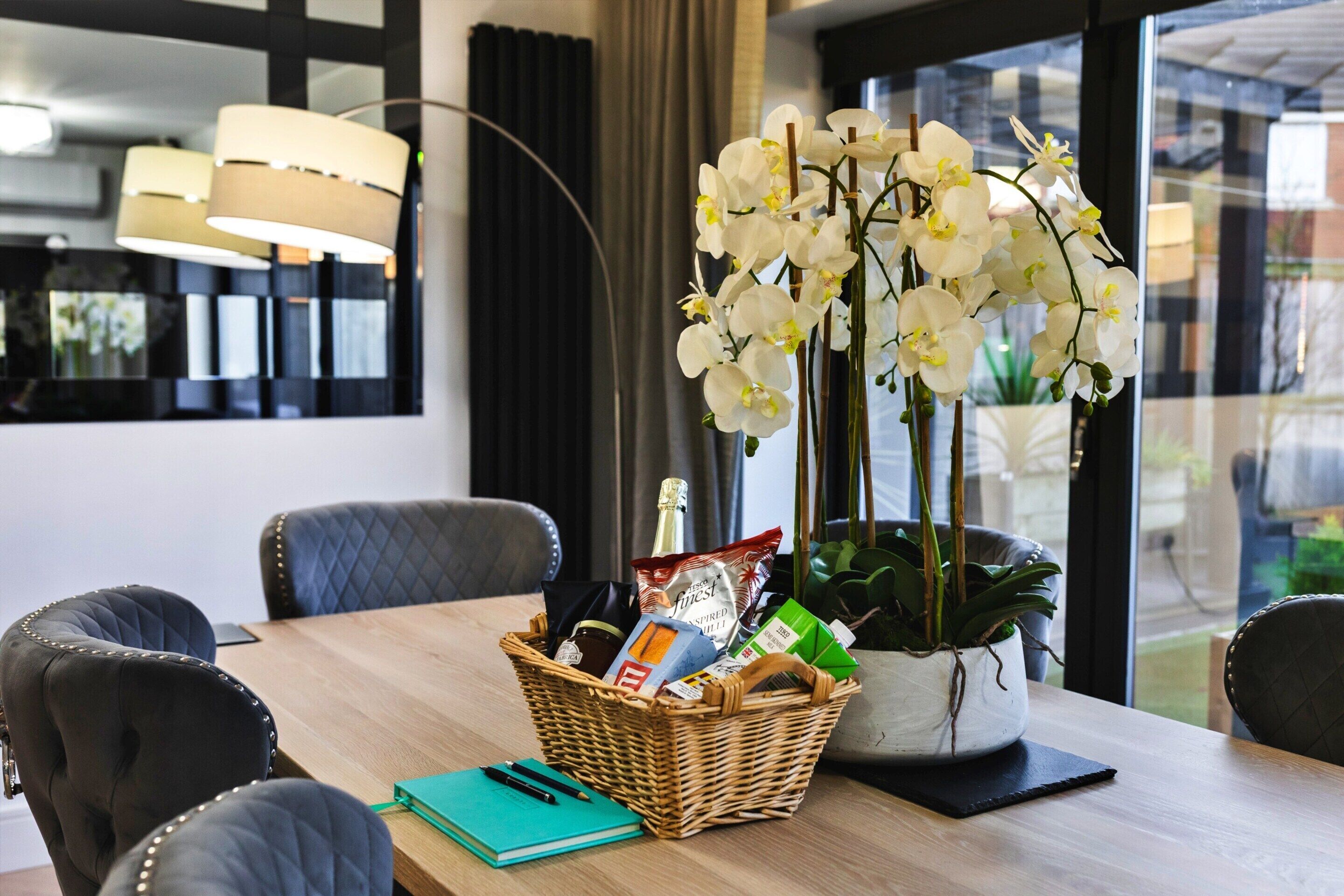 Une chambre d’hôtel avec une table à manger en bois, un panier en osier contenant des en-cas, un carnet, un stylo et une orchidée en pot.