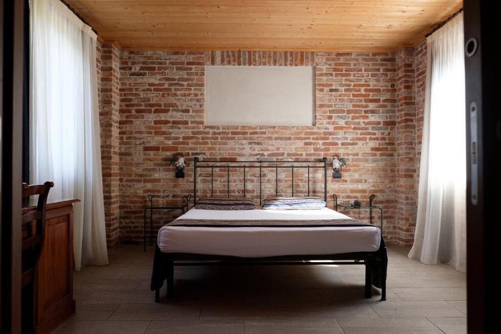 A room with a brick wall, a bed with a metal frame, and curtains on the windows.