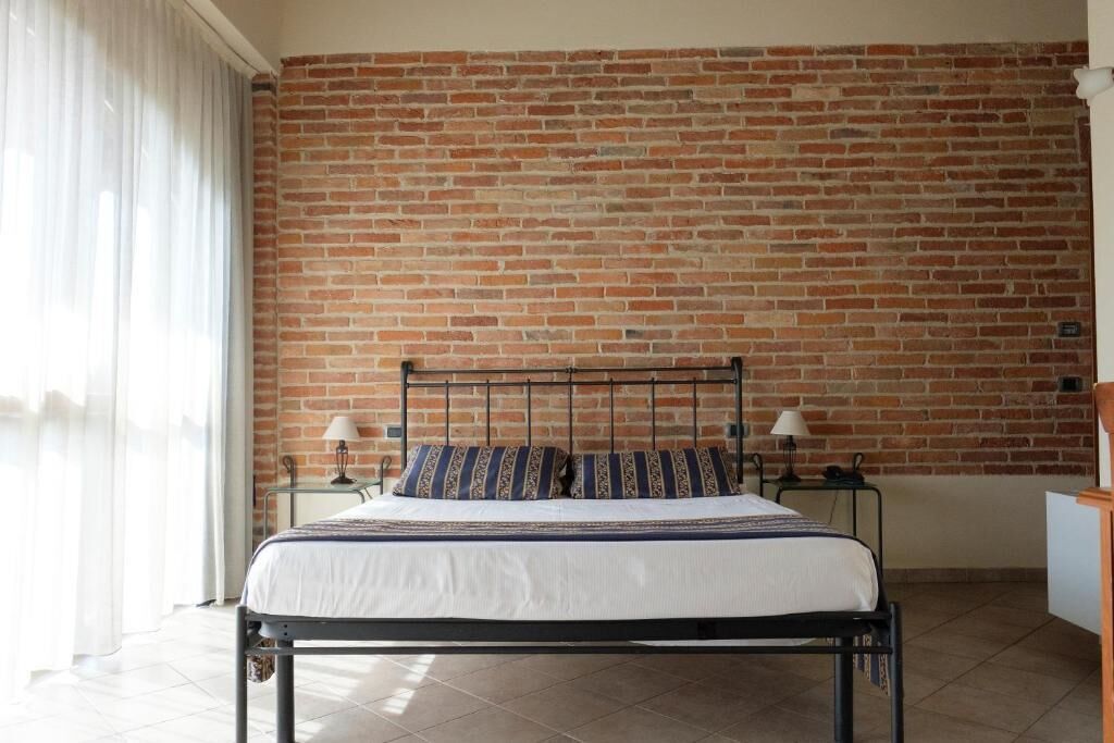 Une chambre avec un mur en briques, un cadre de lit en métal, un couvre-lit blanc, deux tables de chevet avec des lampes et une fenêtre avec des rideaux.