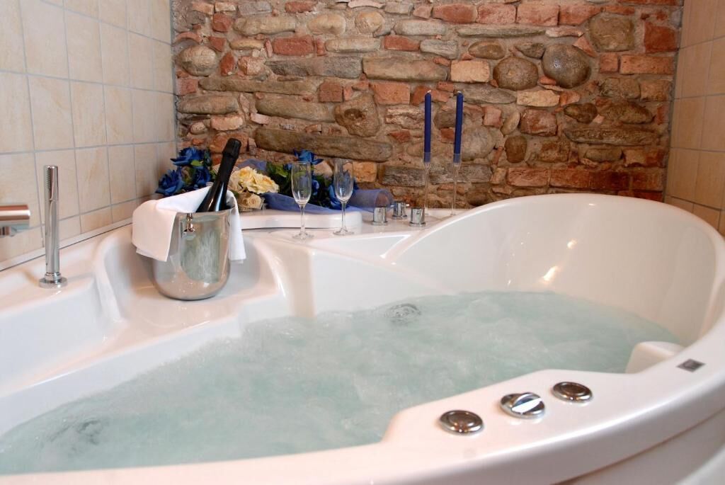 A bathtub with a stone wall backdrop, a glass container with a bottle, and a set of wine glasses.