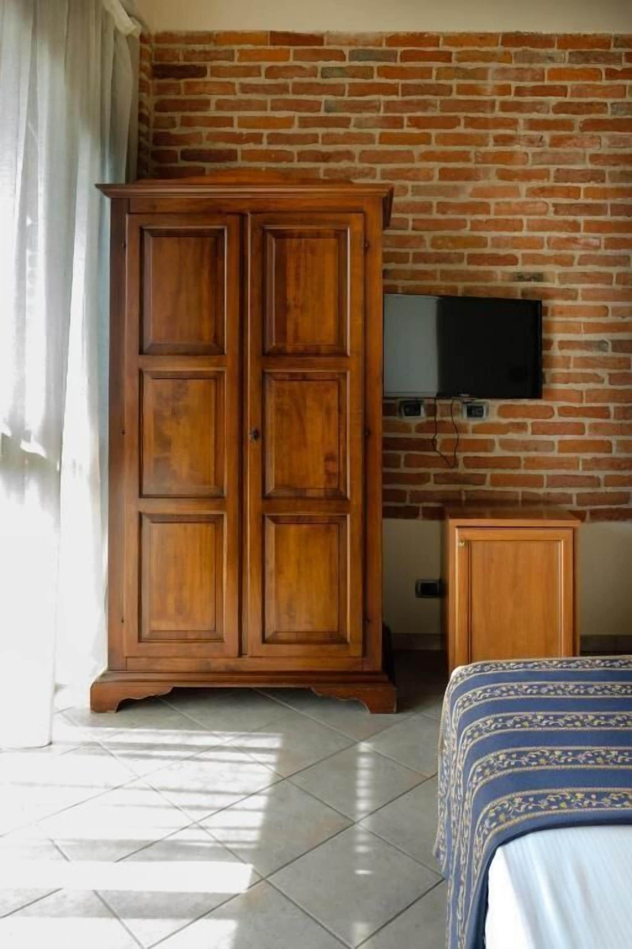 A room with a brick wall, a wooden wardrobe, a television mounted on the wall, and a bed with patterned bedding.