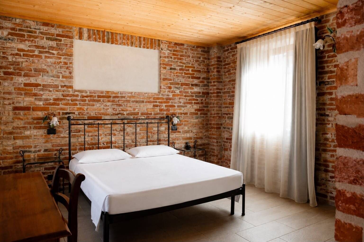Ein Zimmer mit Backsteinwand, einem Bett mit weißer Bettwäsche, einer Holzbalkendecke und einem Fenster mit Vorhängen.