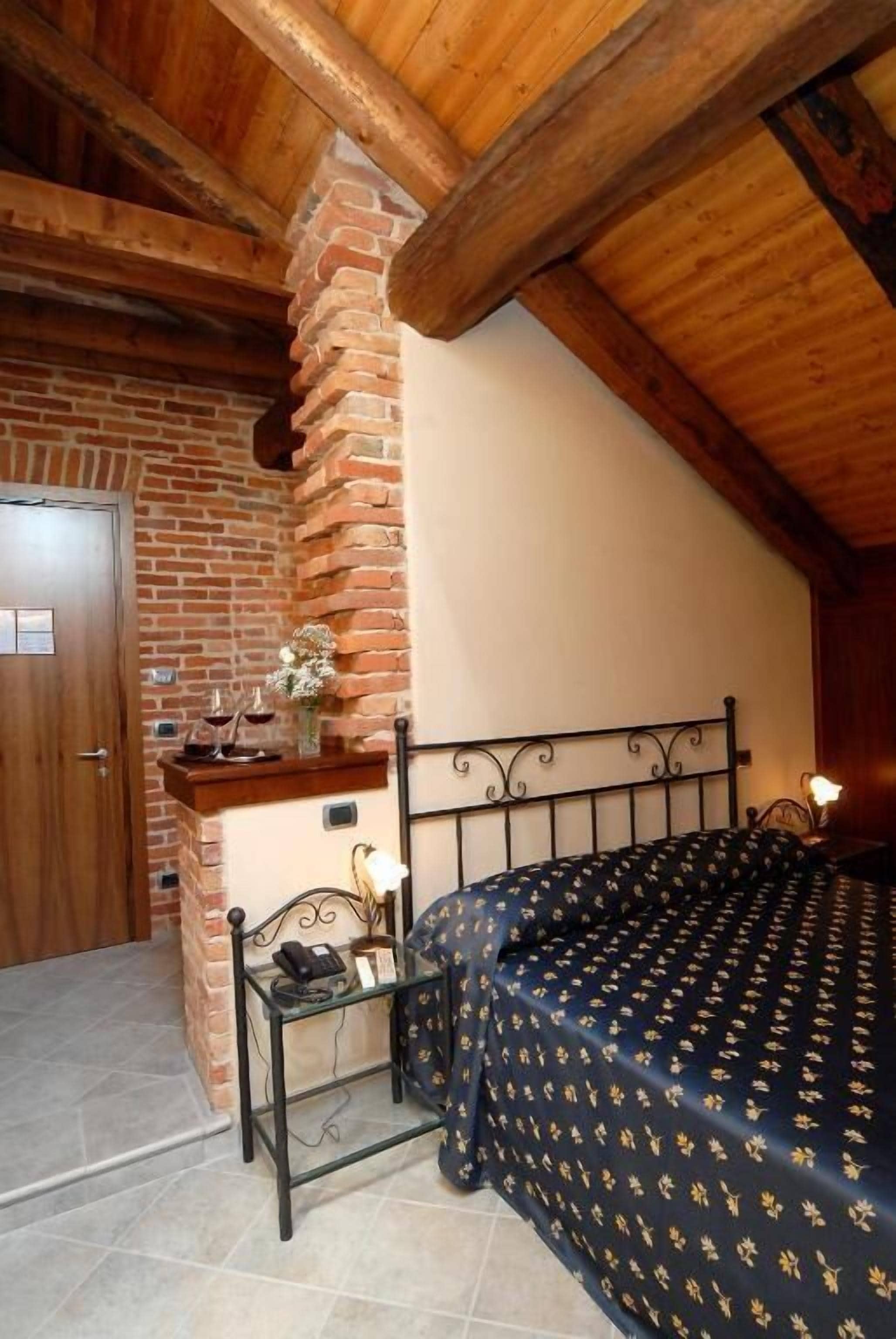 A bedroom with a brick wall, a wooden ceiling, a bed with a patterned bedspread, a bedside table with a lamp, and a small shelf with wine glasses.