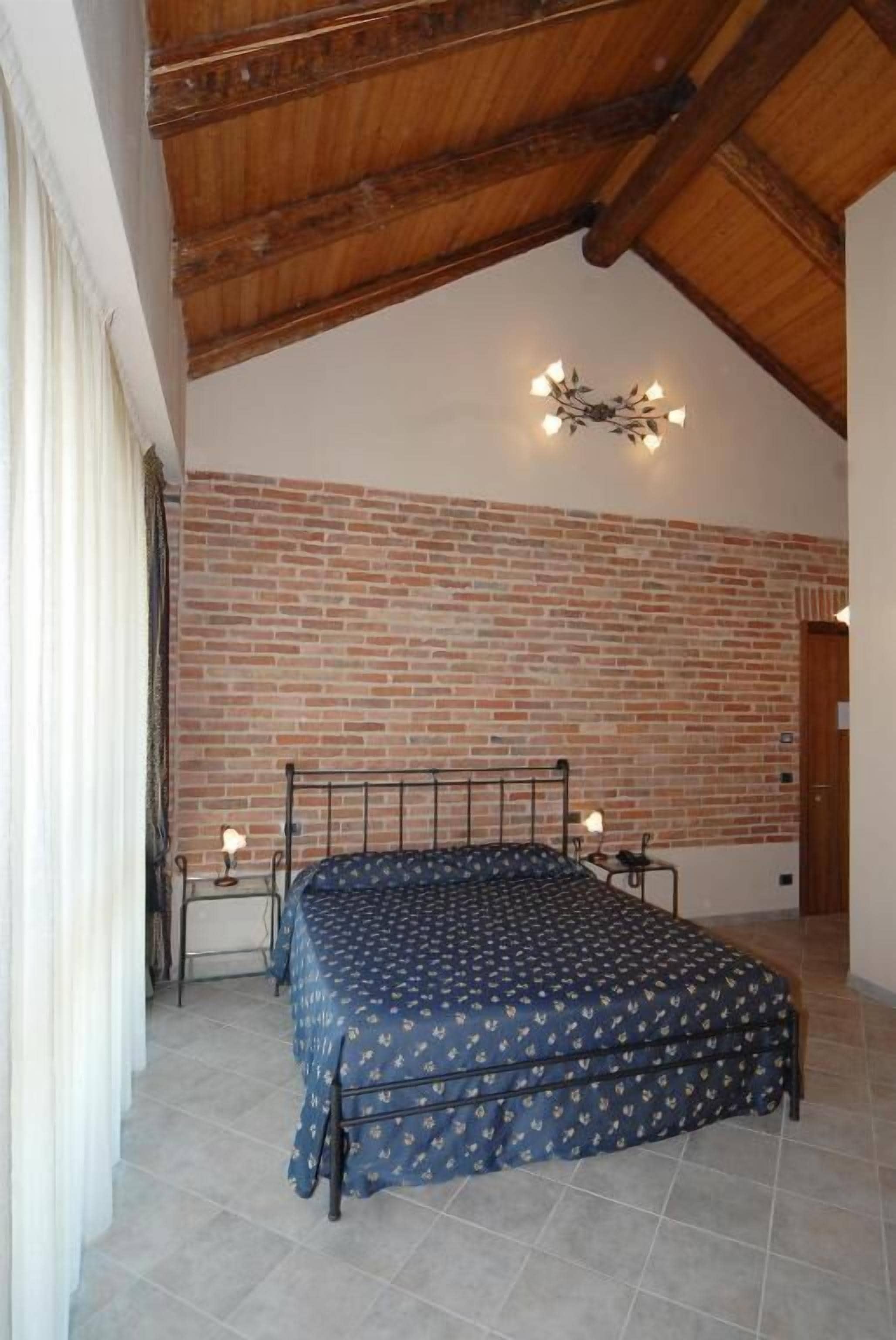 Een slaapkamer met een bakstenen muur, een bed met een geborduurde spreideken, een houten plafond en een wandlamp.