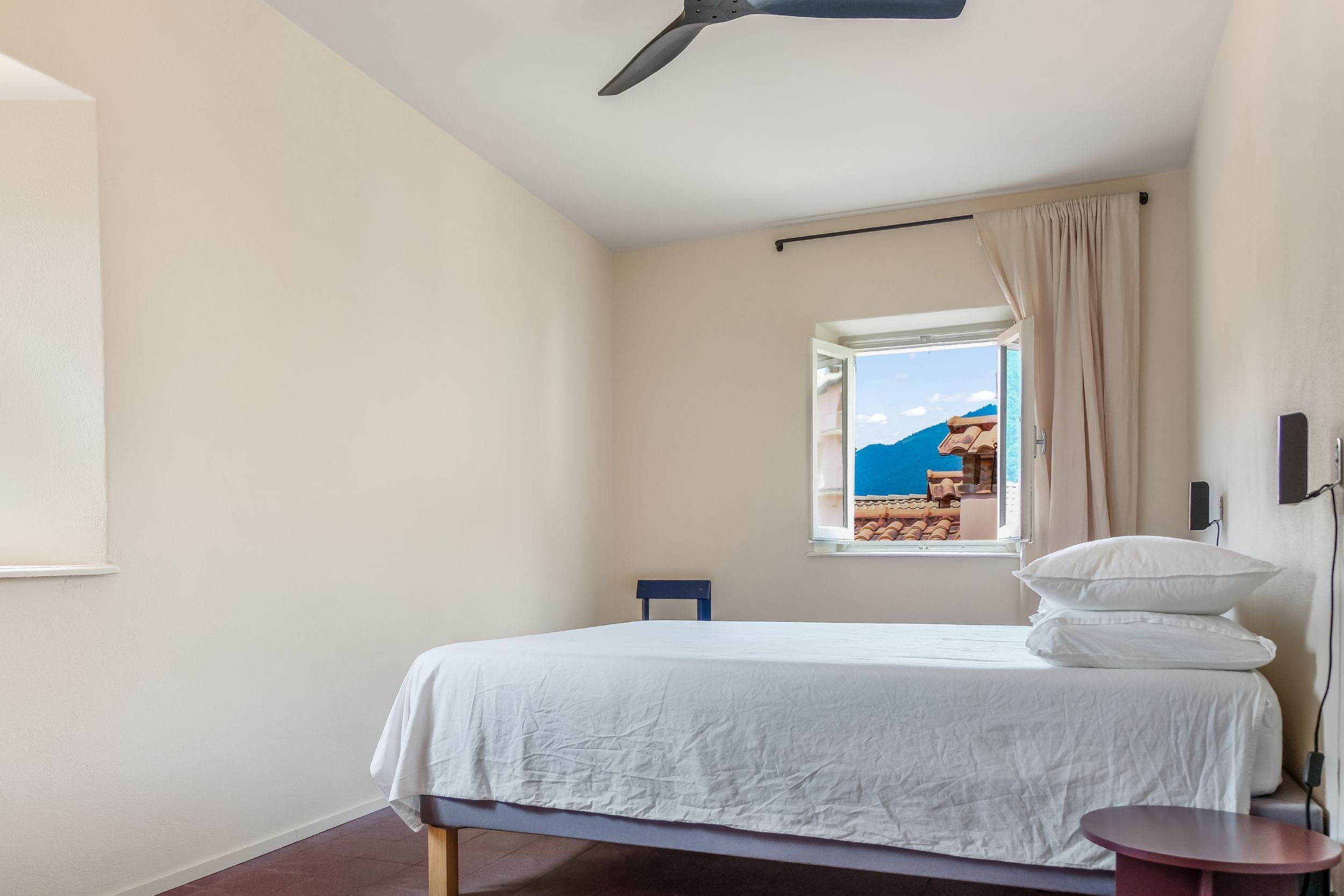 A bedroom with a bed, a window with a view of a mountain and the sea, a ceiling fan, and a bedside table.