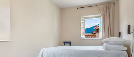 A bedroom with a bed, a window with a view of a mountain and the sea, a ceiling fan, and a bedside table.