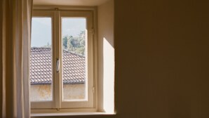 Ein Zimmer mit einem Einzelbett, einem Fenster mit Vorhängen und Blick auf ein Ziegeldach.