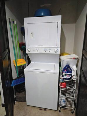 Une petite buanderie équipée d’un lave-linge et d’un sèche-linge, ainsi que d’un balai, de plusieurs brosses et de produits de nettoyage.