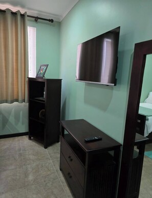 A room with a television mounted on the wall, a small wooden table with a remote control, a dark wooden cabinet, and a window with curtains. - Amazing Oceanfront With Access Crocs Resort (Jacó)