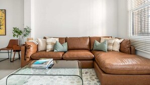 A brown leather sofa with multiple cushions, a glass coffee table, a wooden side table with a vase, and a chair. - 44 Adelaide Street (Surry Hills)