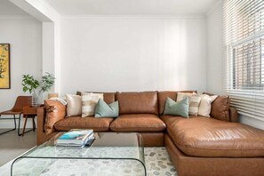 Ein braunes Ledersofa mit mehreren Kissen, ein Glastisch, ein Holznachttisch mit einer Vase und ein Sessel.