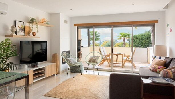 A modern living room with a flat-screen TV, a dining area with a glass table, and a balcony with a view of palm trees. - Albufeira Poolside Family Vila (Albufeira)