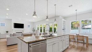 A modern kitchen with a central island, stainless steel appliances, pendant lighting, and a view of the outdoors through large windows. - NEW! Large Luxury Beach Villa: Heated Pool Oasis (Jacksonville Beach)