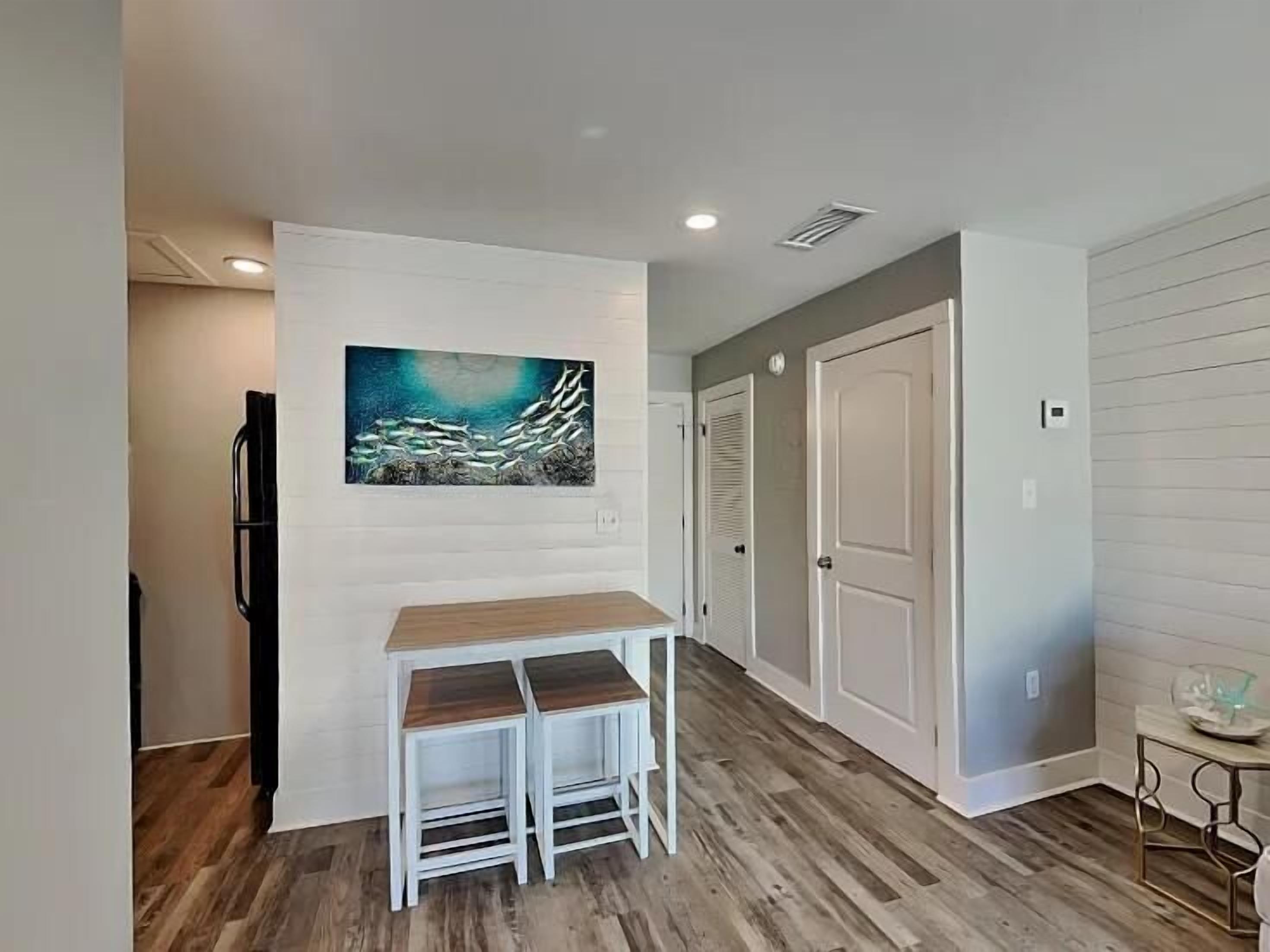 A modern kitchen with a wooden table and two chairs, a painting on the wall, and a refrigerator.