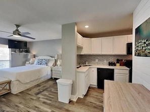 A compact hotel room with a bed, bedside table, lamp, and a small kitchen area with cabinets and a dishwasher. - Sandpiper Cove 8246 (Destin)