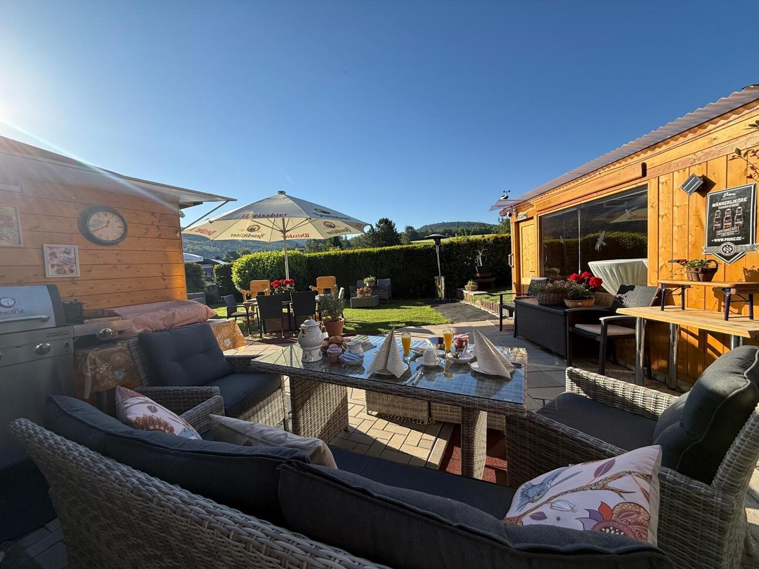 Een terras buiten met rieten meubilair, een barbecue en een houten gebouw met een bord.