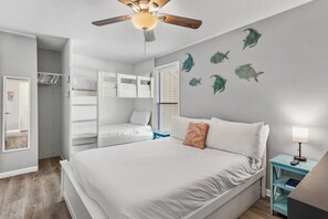 A bedroom with a bed, a ceiling fan, a window with blinds, a wall-mounted shelf, and a door. - Sandpiper Cove 2015 (Destin)