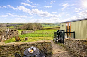A rustic outdoor seating area with stone walls, a table with cups and a basket of bread, and a view of rolling hills in the distance.