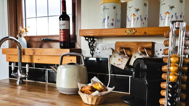Una cocina con una botella de vino, un hervidor, una cafetera, una cesta de pasteles y una tetera sobre la encimera.