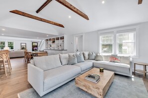 A spacious living area with a light-colored sectional sofa, a wooden coffee table, and a built-in shelving unit. - Elevated Osterville Escape with Heated Pool RPAM128 (Osterville)