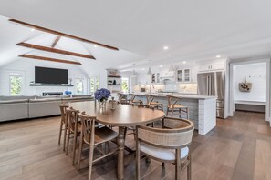 A modern kitchen with white cabinetry, a central island, and a dining area with wooden chairs. - Elevated Osterville Escape with Heated Pool RPAM128 (Osterville)