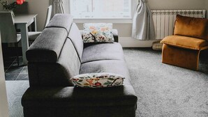 Un salon avec un canapé gris, un fauteuil marron, une table à manger avec des chaises, une fenêtre avec des rideaux et un miroir fixé au mur.