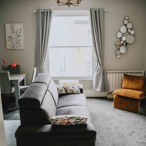 Ein Wohnzimmer mit grauem Sofa, braunem Ohrensessel, Esstisch mit Stühlen, einem Fenster mit Vorhängen und einem wandmontierten Spiegel.