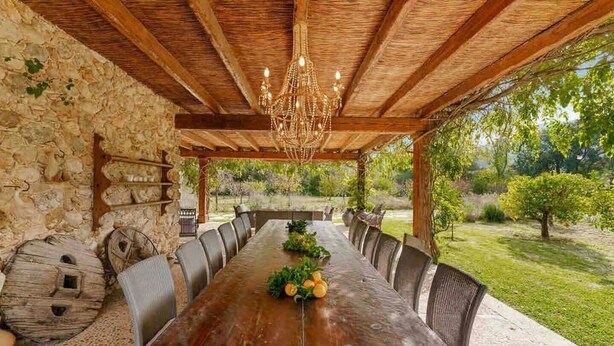 A covered outdoor dining area with a long wooden table, stone walls, and a view of a garden. - The Dream Valley of Mallorca (Pollença)