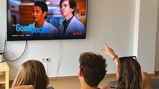 A living room with a flat-screen TV mounted on the wall displaying a show, a sofa, and a person pointing at the screen. - Beachfront villa in Valencia for up to 25 people. (València)