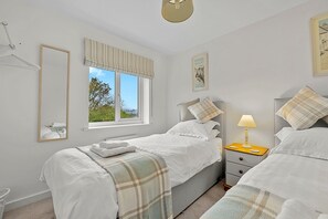 Ein Schlafzimmer mit zwei Betten, einem Nachttisch mit Lampe, einem Fenster mit Vorhängen und einem gerahmten Bild an der Wand.