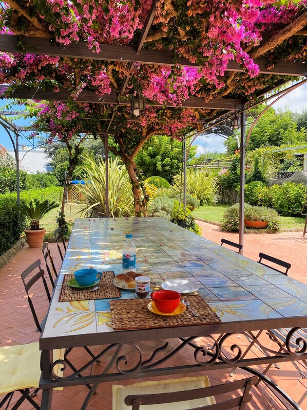 Un espace de restauration en plein air avec une table dressée pour deux personnes, entouré d’une végétation luxuriante et agrémenté d’une pergola couverte de plantes grimpantes en fleurs.