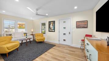A modern living room with a sofa, two armchairs, a coffee table, and a rug. There is a door with a glass panel, a painting on the wall, and a flat-screen TV on a stand.