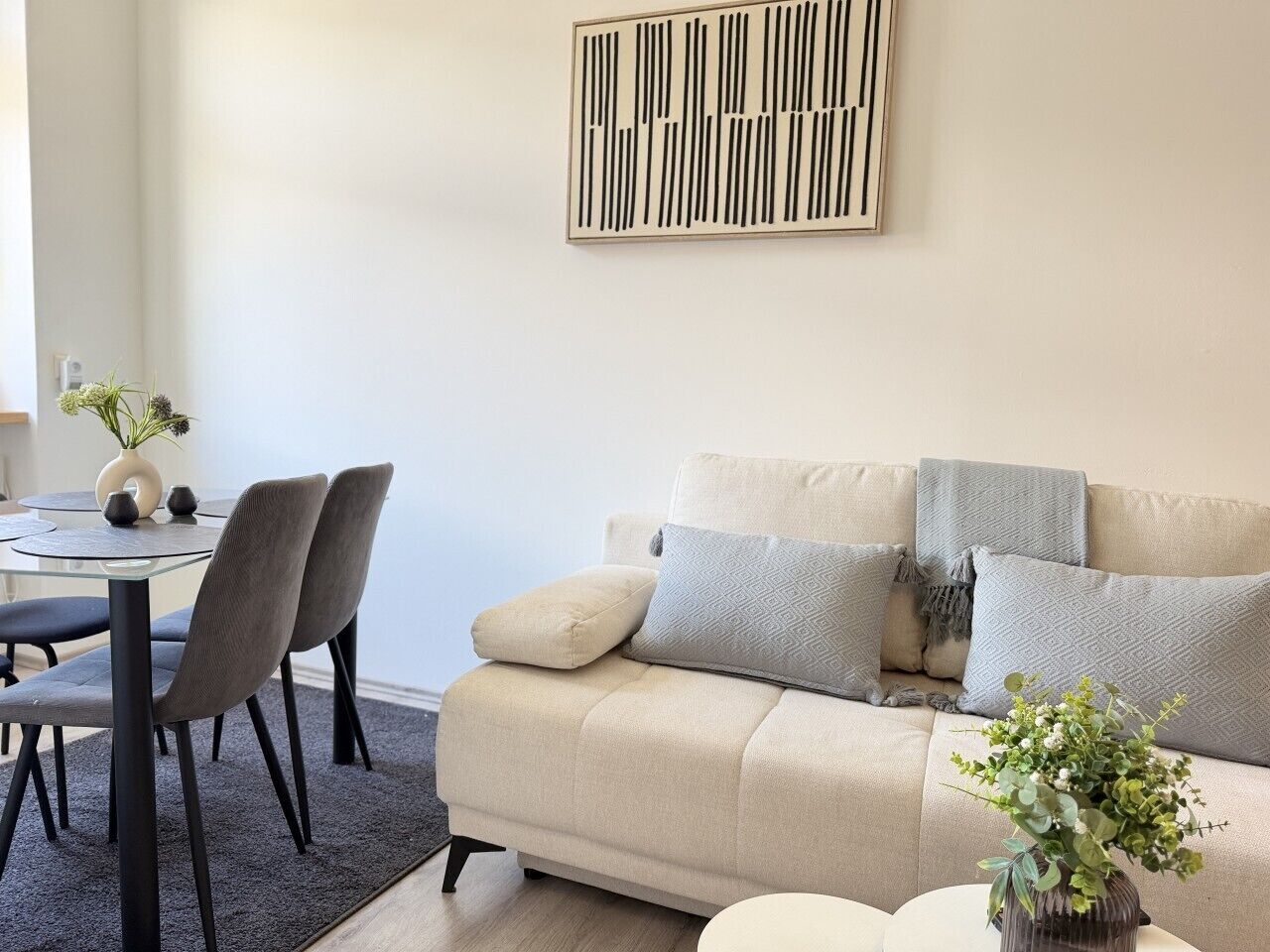 A modern living room with a beige sofa, two grey chairs, a glass coffee table, and a decorative vase with plants.
