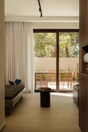 A modern bedroom with a bed, a small table, and a view of an outdoor balcony with chairs and greenery. - Linum Hill Elegant 1BR Apt with Private Pool (Agios Nikolaos)