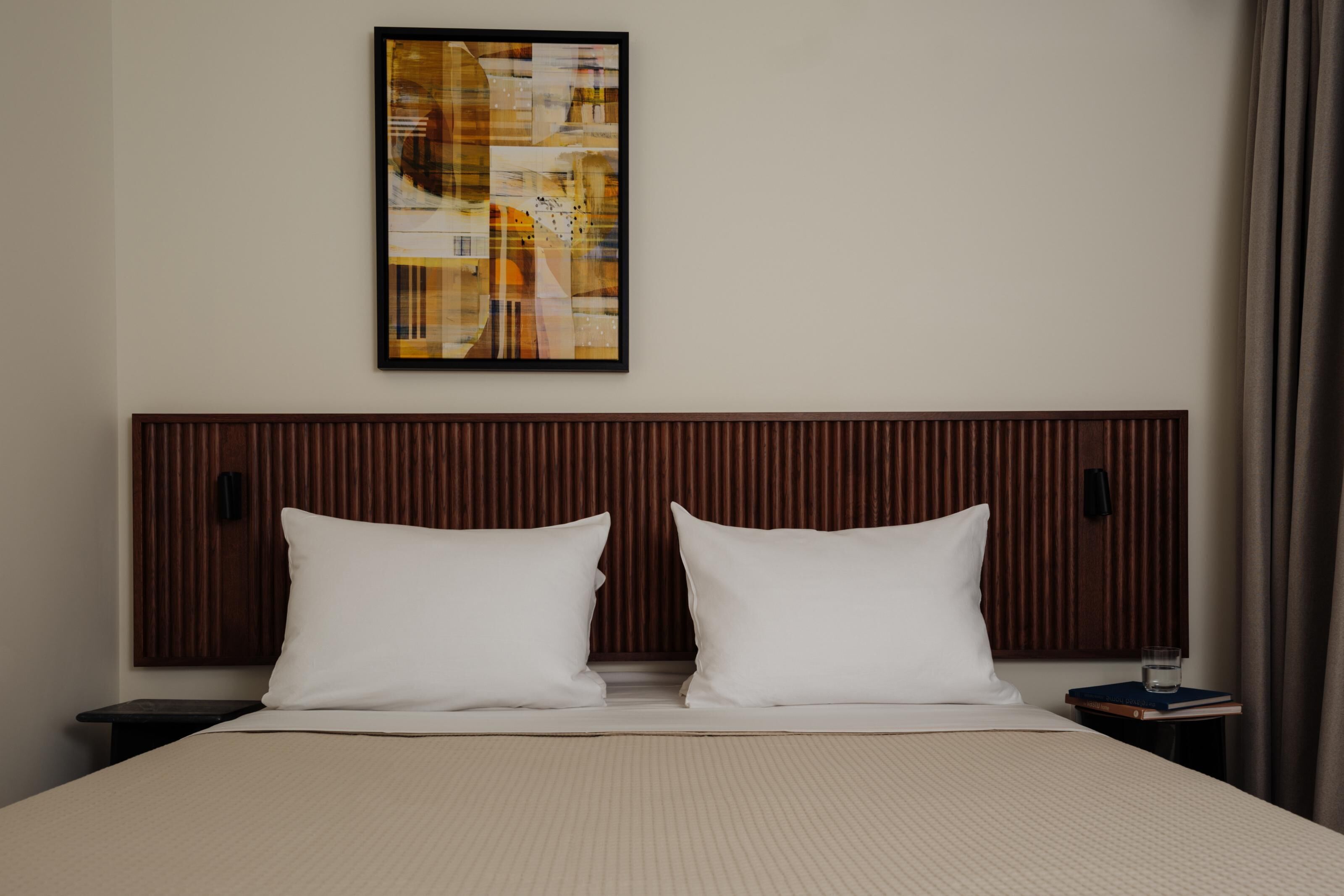 Quarto de hotel com uma cama, duas almofadas, mesa de cabeceira com um livro e um copo, e uma pintura abstrata na parede.