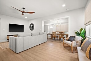 A modern living room with a sectional sofa, a dining area with wooden chairs, a flat-screen TV mounted on the wall, and a ceiling fan. - Steps to the Beach & Bocce Ball (Redington Beach)