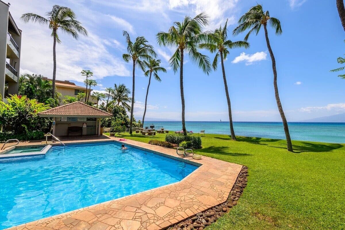 A swimming pool with a tiled deck, surrounded by palm trees and a view of the ocean.
