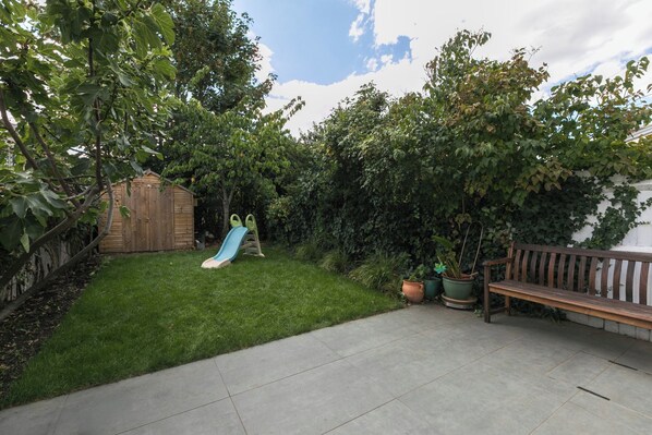 A backyard with a wooden playhouse, a slide, a bench, and potted plants. - Charming 2BDR Garden Home in Turnpike Lane (Greater London)