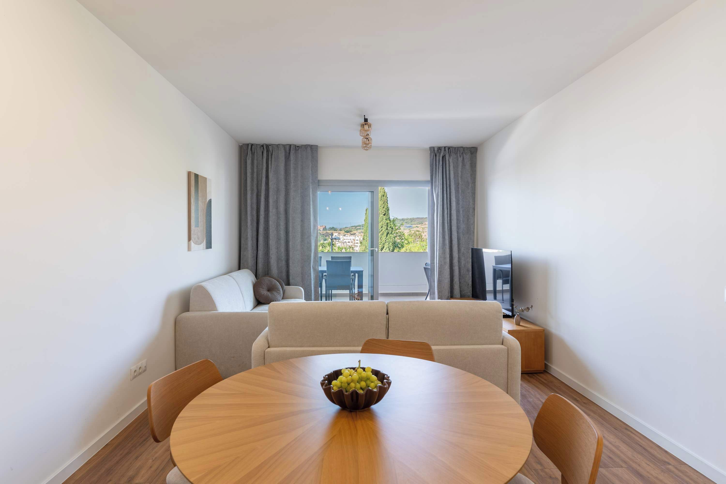 A modern living room with a round wooden dining table, a beige sofa, and a television on a stand.