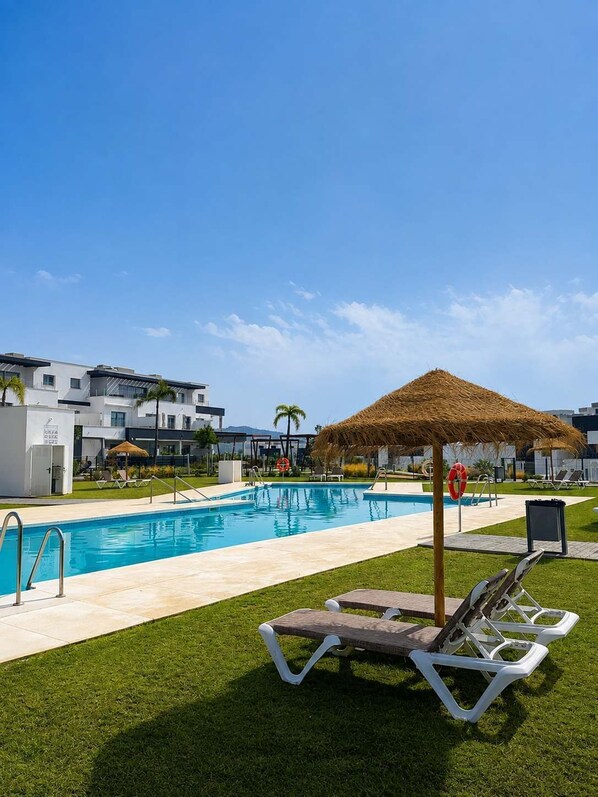 A swimming pool with lounge chairs and a thatched umbrella, surrounded by a well-maintained lawn and modern buildings in the background. - Habitat Valle Romano B | Pool, Gym & Playground (Estepona)