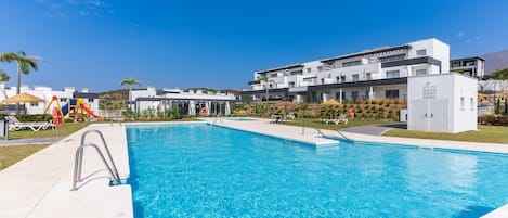 A swimming pool with a clear blue water surface, surrounded by a white building with balconies and a playground area with a slide.