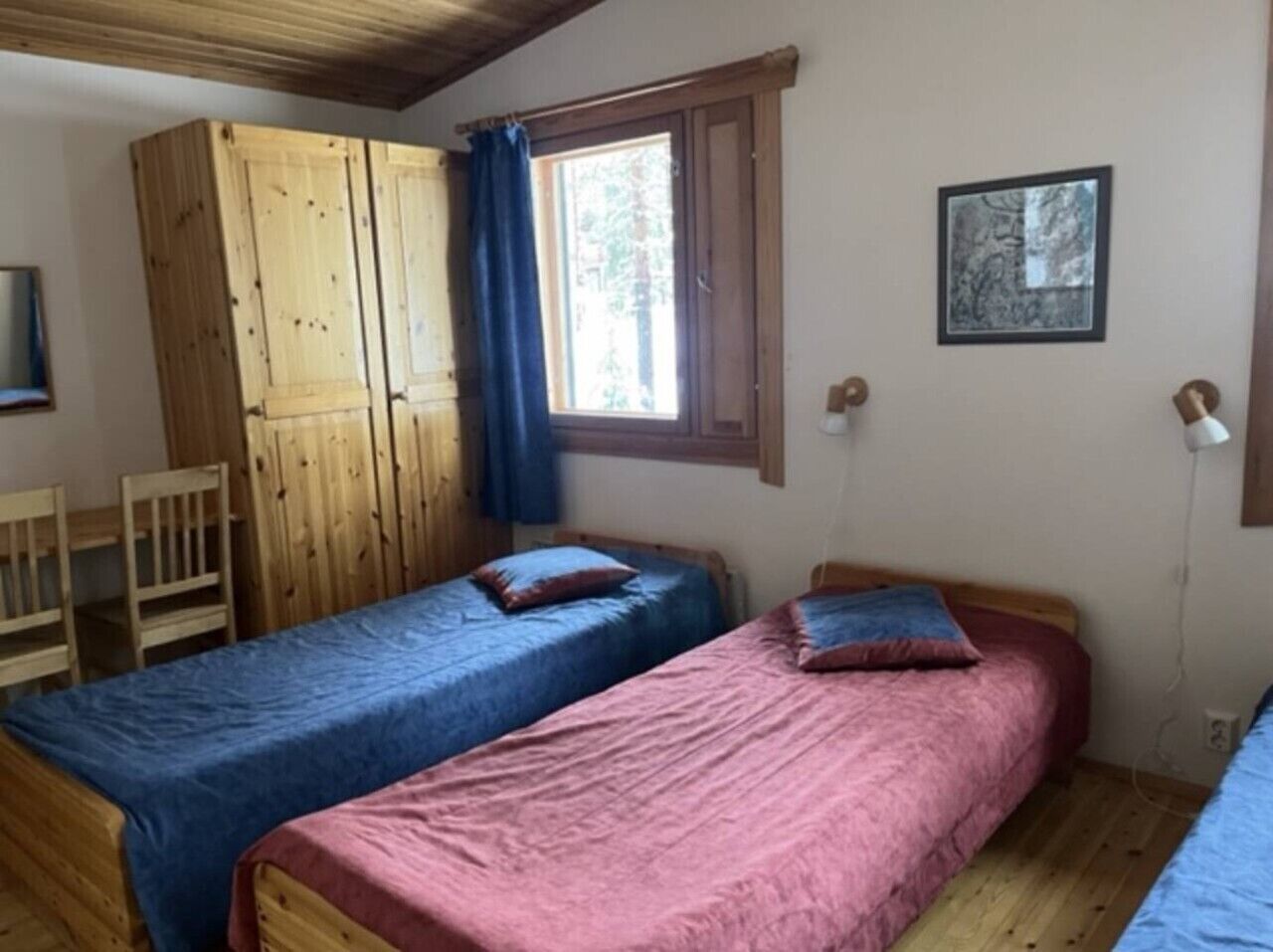 A room with two single beds, a wooden wardrobe, a window with curtains, and a framed picture on the wall.