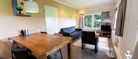 A modern living space with a wooden dining table, a sofa, a TV, and a large window with curtains.