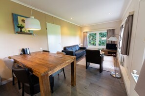 A modern living space with a wooden dining table, a sofa, a TV, and a large window with curtains. - Single-Story Chalet in a Wooded Area Adjacent to a Meadow (Soest)