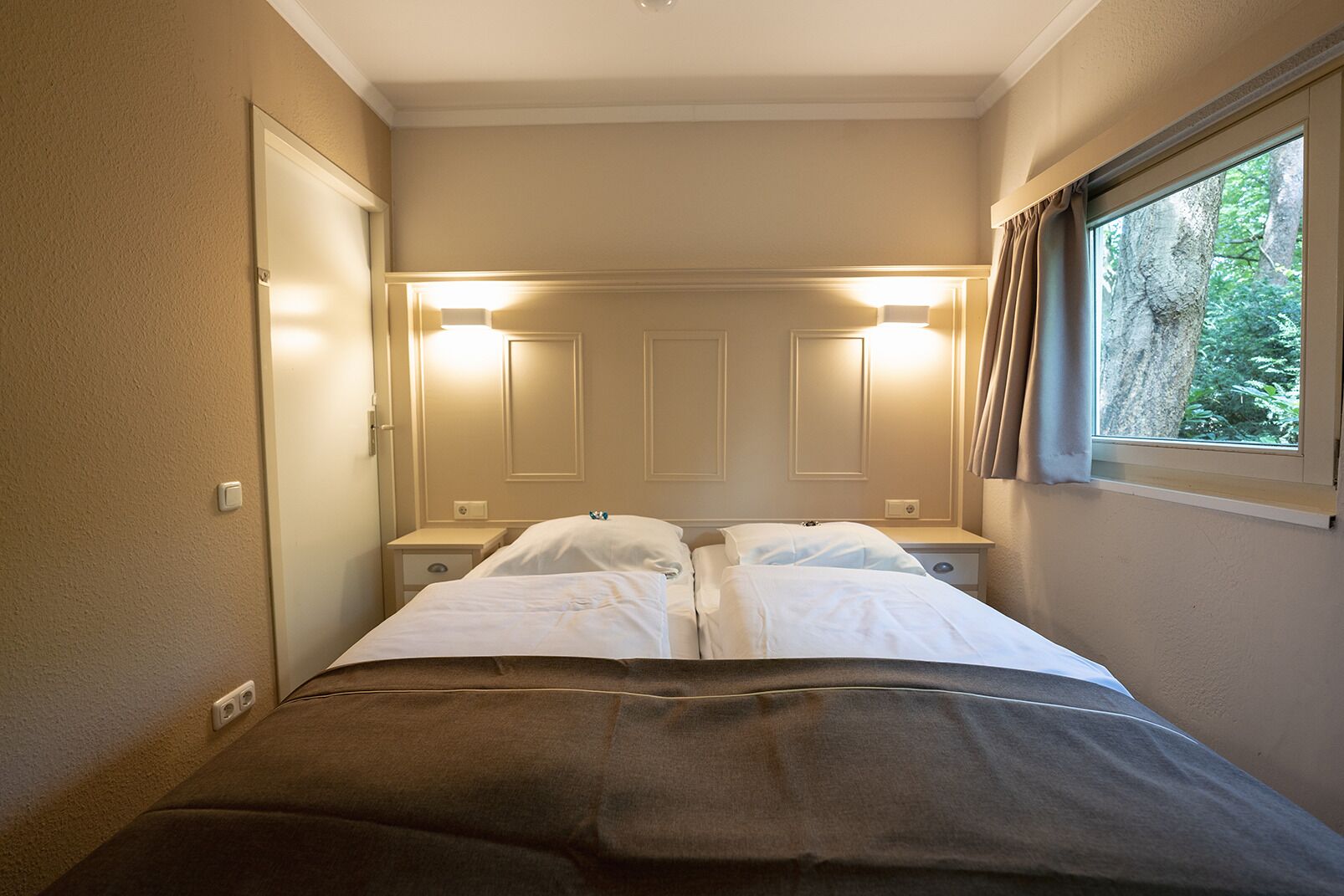 A narrow hotel room with two beds, bedside tables, and a window with curtains.