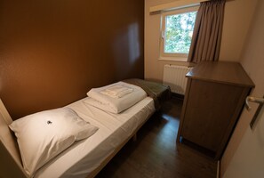 Ein kleines Zimmer mit einem Einzelbett, einem Holzkästen und einem Fenster mit Vorhängen.