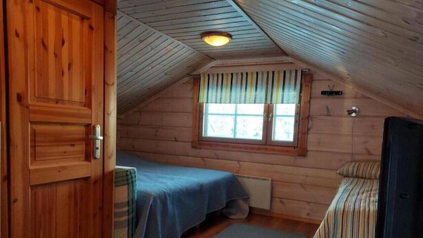 Pieni, yhden huoneen mökki, jossa on puuovi, sinisellä päiväpeitteellä varustettu sänky, verhoilla varustettu ikkuna ja televisio jalustalla.