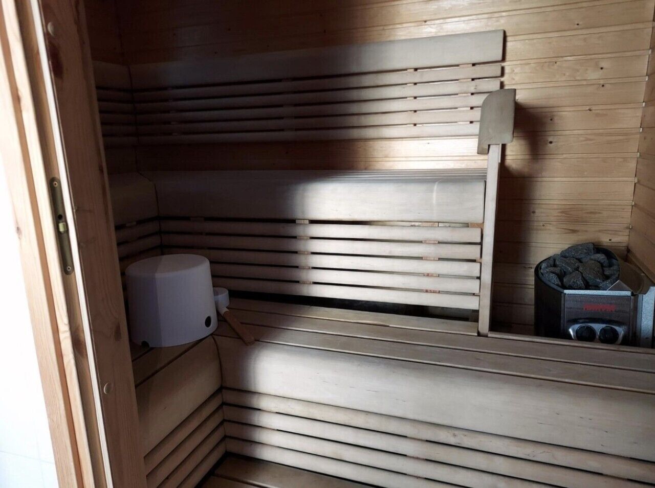 A sauna room with wooden slats, a white heater, and a charcoal container.
